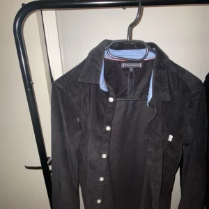 Svart manchesterjacka från Tommy Hilfiger - Snygg svart manchesterjacka från Tommy Hilfiger med knappar framtill. Jackan har en klassisk krage och är perfekt för en stilren look. Den är långärmad och har en diskret logga på baksidan. Perfekt för höst och vår! Storlek 164 ungdom skulle säga att den passar någon runt 165-170 
