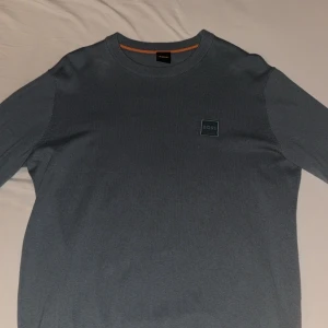 Blå tröja från BOSS - Säljer en stilren blå tröja från BOSS med rund hals och ett diskret märke på bröstet. Tröjan har långa ärmar och är perfekt för en avslappnad look. Passar bra till både jeans och chinos. 