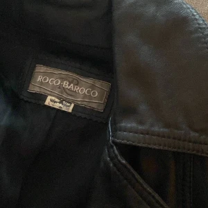 Svart skinnrock från Roco Baroco äkta skinn - Säljer en svart skinnrock från Roco Baroco. Rocken har en klassisk design med långa ärmar och knappar framtill. Den har stora fickor och en stilren krage. Perfekt för en edgy look! Nypris 10 000kr