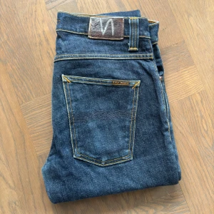 Mörkblåa Nudie Jeans - Säljer ett par mörkblåa Nudie Jeans i modellen "Grim Tim", storlek W32 L34. Tillverkade i 100% bomull med en rak passform, vilket ger en stilren och bekväm look. Jeansen är i ett bra skick och är i väldig bra kvalitiet. Nypris 1600 kr.