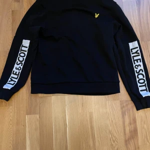 Svart tröja från Lyle & Scott - Säljer en svart tröja från Lyle & Scott med deras logga på bröstet och tryck ärmarna . Tröjan har en avslappnad passform och rund halsringning. Perfekt för en stilren look. Storlek S men passar garanterat M (~175cm-180cm).
