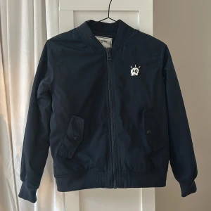 Mörkblå bomberjacka från Zadig & Voltaire - Snygg mörkblå bomberjacka från Zadig & Voltaire med vit text på ryggen. Jackan har dragkedja framtill och två fickor med knappar. Perfekt för en stilren look.