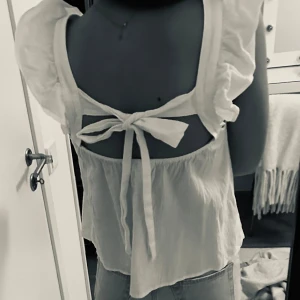 Vit blus med volangdetaljer - Charmig vit blus med volangdetaljer och knytning i ryggen. Perfekt för en somrig look med sina ärmlösa axlar och luftiga design.