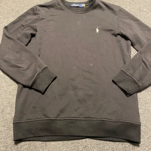 Svart tröja från Polo Ralph Lauren - Säljer en stilren svart tröja från Polo Ralph Lauren med deras ikoniska logga broderad i vitt på bröstet. Tröjan har långa ärmar och en klassisk rund halsringning. Perfekt för en avslappnad och snygg look.