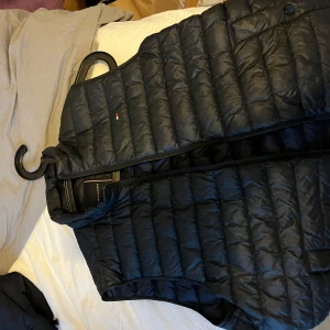 Svart quiltad dunväst från Tommy Hilfiger - Säljer en stilren svart quiltad dunväst från Tommy Hilfiger. Västen har en diskret logga på bröstet och dragkedja framtill. Perfekt för lager-på-lager under kyligare dagar.