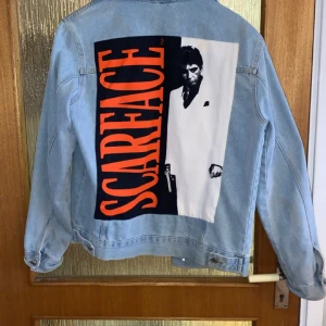 Ljusblå jeansjacka från Pull&Bear - Cool ljusblå jeansjacka från Pull&Bear med Scarface-tryck på ryggen och bröstet. Jackan har knappar framtill och två bröstfickor med lock. Riktigt unik jacka i bra skick. Köpt för ungefär 850kr