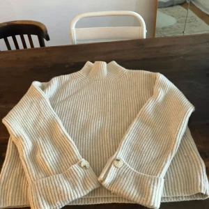 Beige ribbad tröja med knappar - Säljer en stilren beige ribbad tröja med hög krage och långa ärmar. Tröjan har dekorativa knappar vid ärmsluten som ger en extra touch. Perfekt för kyligare dagar. Något nopprig men ska kunna tas bort med ex ullkam 
