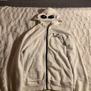 Vit huvtröja med glasögon från C.P. Company - Säljer en unik vit huvtröja från C.P. Company med inbyggda glasögon i huvan. Tröjan har dragkedja framtill och två fickor. Perfekt för en stilren och modern look. Tröjan är riktigt och QR koden kan skannas!!
