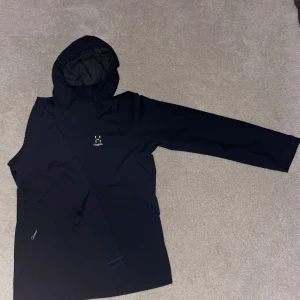 Svart regnjacka från Haglöfs - Säljer en stilren svart regnjacka från Haglöfs med huva och långärmad design. Jackan är tillverkad i ett vattentätt material med PROOF-teknologi, perfekt för regniga dagar. Den har en minimalistisk look med diskret logga på bröstet.