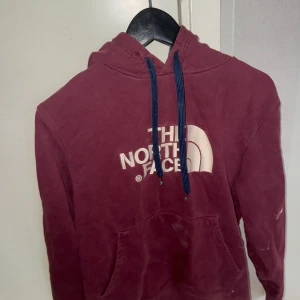 Vinröd hoodie från The North Face - Säljer en vinröd hoodie från The North Face med vit logga på bröstet. Den har en justerbar huva med blå snören och en praktisk magficka. Perfekt för kyliga dagar!