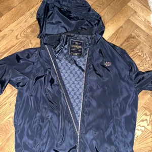Mörkblå vindjacka från Morris - Snygg mörkblå vindjacka från Morris med dragkedja och huva. Jackan har ett stilrent mönster på insidan och en liten broderad detalj på bröstet. Perfekt för blåsiga dagar.