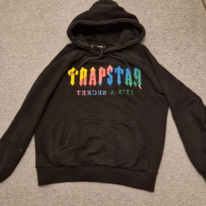 Svart hoodie från Trapstar - Säljer en svart hoodie från Trapstar med färgglad text på framsidan. Hoodien har en klassisk design med känguruficka och justerbar huva. Perfekt för en avslappnad stil.