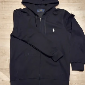 Svart hoodie från Polo Ralph Lauren - Snygg svart hoodie från Polo Ralph Lauren med dragkedja och klassisk logga på bröstet. Perfekt för en avslappnad stil. Den har långa ärmar och en praktisk huva med dragsko.
