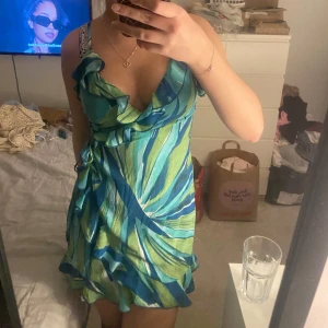 Grönblå omlottklänning - Snygg grönblå omlottklänning med volangdetaljer och knytning i midjan. Klänningen har ett livfullt mönster i olika nyanser av grönt och blått, perfekt för sommaren😍🩵💚💙Den är väldigt fin på och justerbar i passformen då man knyter den! 