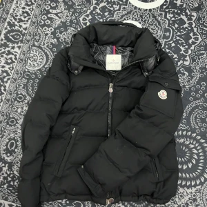 Moncler montgenevre - Säljer en snygg svart dunjacka från Moncler med dragkedja och broderad logga på ärmen. Jackan har en quiltad design och är perfekt för kyligare väder. Den har praktiska fickor med dragkedjor och en hög krage för extra värme. Passar perfekt för vinterns kalla dagar. 