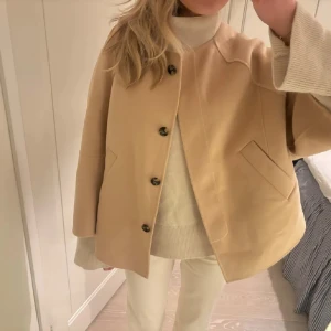Beige trekvartsärmad kappa med knappar - Säljer en stilren beige kappa med knappar framtill. Jackan har en klassisk design med långa ärmar och två fickor. Perfekt för att hålla stilen i kyligare väder.