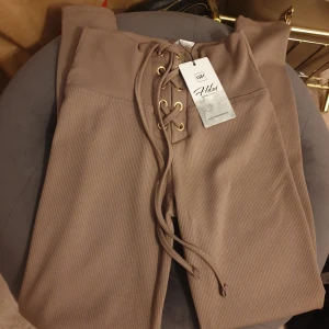 Bruna ribbade byxor med snörning - Helt nya!! Snygga bruna ribbade byxor från Hedni med hög midja och snörning framtill. Perfekta för en stilren look med en touch av detaljrikedom. Byxorna har en tight passform som framhäver figuren. 