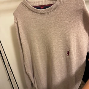 Beige tröja från Polo Ralph Lauren - Säljer en stilren beige tröja från Polo Ralph Lauren med det klassiska broderade logotypen i rött på bröstet. Tröjan har en rund halsringning och långa ärmar, perfekt för en avslappnad och elegant look.
