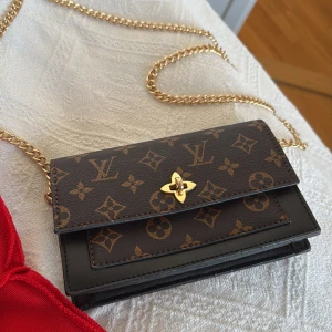 Brun monogram axelväska med guldkedja - Snygg brun axelväska med klassiskt monogrammönster. Väskan har en elegant guldkedja och ett stilrent spänne framtill. Perfekt för att ge din outfit en lyxig touch.