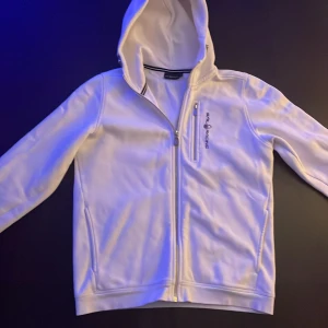 Vit hoodie från Sail Racing - Säljer en stilren vit hoodie från Sail Racing med dragkedja och huva. Den har en bröstficka med dragkedja och märkeslogga på bröstet. Perfekt för en avslappnad stil.