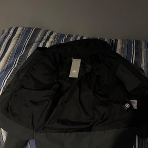 Svart pufferjacka med huva - Snygg grå canada goose jacka. Jackan har en dragkedja framtill och är perfekt för kyligare väder. Den har en stilren design med en diskret logga på ärmen. Helt ny och är inte använd en enda gång. För mer detalj bilder skriv gärna så svarar jag. Vid snabb affär kan pris diskuteras.