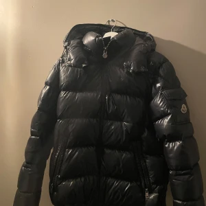 Svart dunjacka från Moncler - Säljer en svart dunjacka från Moncler med en glansig finish. Jackan har en dragkedja framtill och en ficka med Moncler-logga på ärmen. Perfekt för kalla dagar. - lite små hål vid fickorna som man knappt ser och ett på armen som man bara ser själv när man kollar nära, tveka inte på att fråga någon fråga 