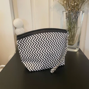 Svartvit mönstrad necessär - Snygg och praktisk necessär med ett svartvitt sicksackmönster. Den har en dragkedja upptill och en liten rem för enkel hantering. 17cm hög, 22cm lång, 8cm djup