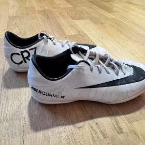Vita Nike Mercurial X fotbollsskor - Snygga vita Nike Mercurial X fotbollsskor med svart Nike-logga och CR7-detaljer. Skorna har snörning och en mönstrad sula för bra grepp. Perfekta för inomhusfotboll.