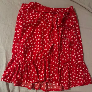 Röd blommig kjol med volang - Charmig röd kjol med vita blommor och volangdetalj. Kjolen har en omlott-design och knytband i midjan för en justerbar passform. Perfekt för en somrig look!