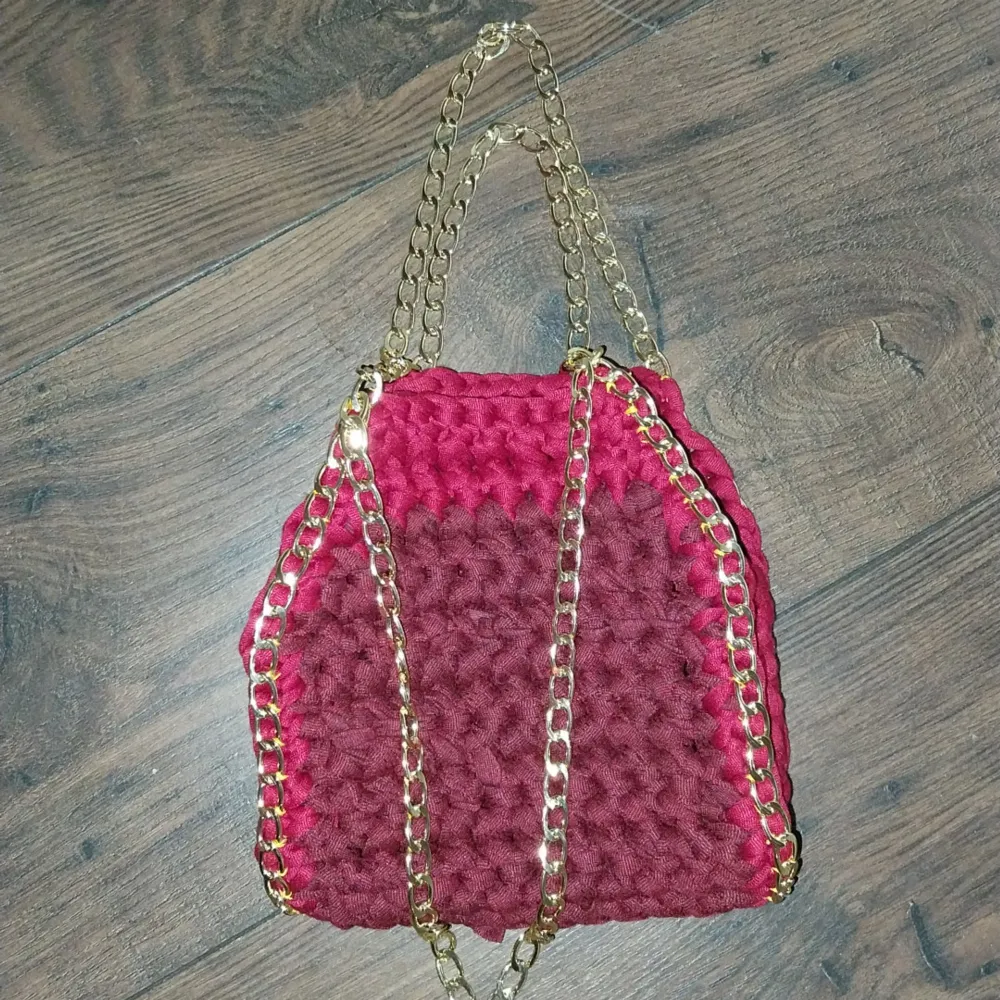 Snygg vinröd virkad väska med guldiga kedjedetaljer. Väskan har en unik textur och är perfekt för att ge din outfit en färgklick. Den har en praktisk storlek och kan bäras i handen.. Laukut & Käsilaukut.