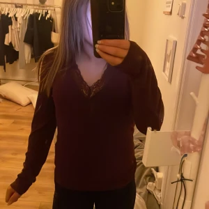 Vinröd långärmad topp med spetsdetalj - Säljer en vinröd långärmad topp med v-ringning och spetsdetalj vid halsen. Perfekt för en stilren och elegant look. Passar bra till både jeans och kjol.