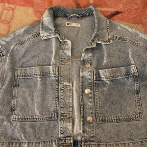 Jeansjacka från Uniqlo - Säljer en klassisk jeansjacka från Uniqlo i ljusblå denim. Jackan har en avslappnad passform med två stora bröstfickor och knappar framtill. Perfekt för en casual look!