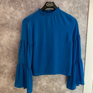 Blå blus från Gina Tricot - Snygg blå blus från Gina Tricot med långa volangärmar och en liten volangdetalj vid halsen.