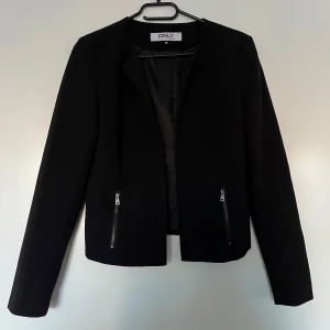 Kavaj, Only, 36, svart - Svart kort kavaj från Only i storlek 36, svart. Kavajen är i nyskick.  Enkel att matcha med alla dina garderobfavoriter för alla tillfällen.  Kort modell. Normal passform. 2 framfickor fram med blixtlås. 100% polyester.  Mått:  Längd ca 56 cm Byst ca 43 cm Midja ca 40 cm Ärmlängd mätt från axeln ca 62 cm