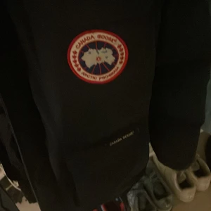 Mörk blå dunjacka från Canada Goose - Säljer en mörk blå dunjacka från Canada Goose med pälsdetalj på huvan. Jackan har en klassisk design med lång ärm och är perfekt för kalla vinterdagar. Den har en tydlig logotyp på bröstet och är tillverkad i högkvalitativa material.