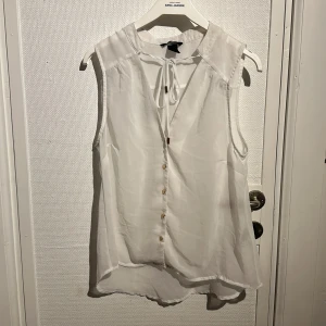 Vit ärmlös blus från H&M - Säljer en stilren vit ärmlös blus från H&M. Blusen har en knytning vid halsen och knappar framtill. Perfekt för en somrig look eller att bära under en kavaj.