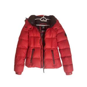 Snygg röd dunjacka från Superdry med dragkedja och huva. Jackan har långa ärmar och praktiska fickor med dragkedjor. Perfekt för kyliga dagar när du vill hålla stilen.