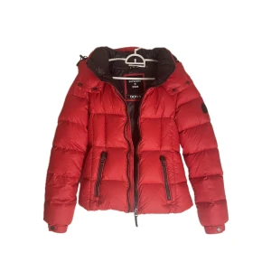 Superdry jacka - Snygg röd dunjacka från Superdry med dragkedja och huva. Jackan har långa ärmar och praktiska fickor med dragkedjor. Perfekt för kyliga dagar när du vill hålla stilen.