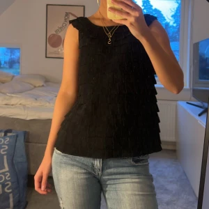 Svart topp - Snygg svart ärmlös topp med volanger i lager-på-lager stil. Perfekt för en stilren look. Toppen har en rund halsringning och är tillverkad i ett mjukt material.