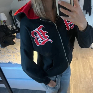 Svart hoodie med röd, svartf - Säljer en cool svart hoodie från Shein med röd insida i luvan och stora bokstäver i rött och vitt på framsidan. Den har dragkedja och är perfekt för en avslappnad stil. Passar bra till jeans eller leggings. Perfekt för höst och vår!