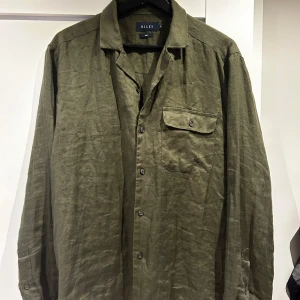 linneskjorta Riley - Säljer en snygg olivgrön skjorta i linne från Riley. Perfekt för både vardag och fest! Skjortan har långa ärmar och en klassisk krage med knappar hela vägen ner. Den har också en praktisk bröstficka med knapp. Passar perfekt till vår och sommar.