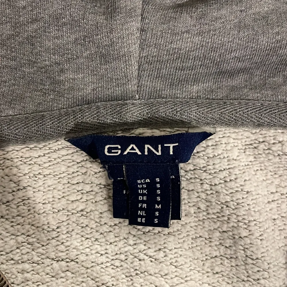 Säljer en snygg grå hoodie från GANT i storlek S. Den har en dragkedja framtill och stora fickor. Perfekt för höst och vår, med ribbade muddar och en mysig huva. GANT-loggan är broderad på bröstet och lite nopprig. Passar både till vardags och chillkvällar. 🩶. Tröjor & Koftor.