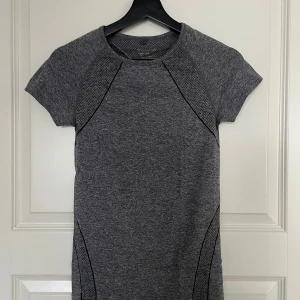 Grå tränings t-shirt  - Snygg grå tränings t-shirt i mycket bra skick. Den har en tight passform och korta ärmar, perfekt för träning. Detaljerna med svarta linjer ger en sportig touch. Materialet är mjukt och stretchigt, vilket gör den bekväm att bära hela dagen.