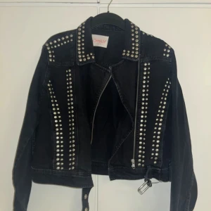 Svart jeansjacka med nitar från ONLY - Säljer en svart jeansjacka från ONLY i storlek M. Jackan har coola nitar på kragen och längs framsidan, vilket ger den en edgy look. Den är gjord av återvunnen bomull och har en bälte i midjan. Perfekt för höst och vår!