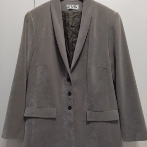 Grå kavaj från Almia - Säljer en stilren gråbeige kavaj från Almia i storlek C44. Kavajen har en klassisk design med tre knappar framtill och långa ärmar. Perfekt för både formella och vardagliga tillfällen. Den är i mycket bra skick och redo att bäras till nästa event!