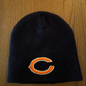 New Era Mössa - Säljer en svart mössa med en broderad orange och vit logotyp på framsidan. Perfekt för att hålla sig varm under kalla dagar. Mössan är i bra skick och passar de flesta huvudstorlekar.