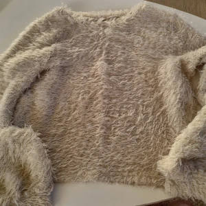 Beige fluffig tröja från Gina Tricot - Mysig och superfluffig beige/vit  tröja från Gina Tricot i storlek XS. Perfekt för kyliga dagar när du vill känna dig både varm och stilren. Tröjan har långa ärmar och en lös passform som ger en avslappnad look. Passar perfekt till jeans eller leggings!