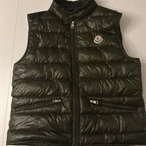 Säljer en snygg svart dunväst från Moncler i mycket bra skick. Västen har en dragkedja framtill och två praktiska fickor med dragkedjor. Perfekt för höst och vår när du vill hålla dig varm men ändå se stilren ut. Passar både till vardags och fest! Har kvitto på den