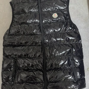 Svart glansig dunväst från Moncler - Säljer en snygg svart glansig dunväst från Moncler. Västen har en dragkedja framtill och två praktiska fickor med dragkedjor. Perfekt för kyliga dagar när du vill hålla stilen. Den är i mycket bra skick och passar perfekt för höst och vår.