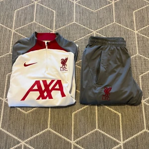 Nike Tracksuit - Säljer en snygg Nike tracksuit i vitt och grått med röda detaljer. Tröjan har en halv dragkedja och långa ärmar, perfekt för fotbollsträning. Byxorna är grå med elastisk midja och röda loggor. Både tröjan och byxorna har LFC-loggan. Perfekt för alla Liverpool-fans! 